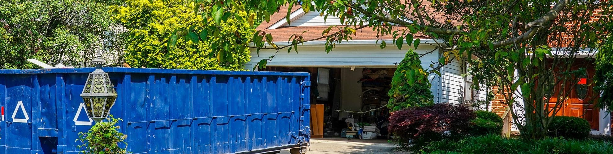 An empty blue dumpster in the driveway of a house with its garage door